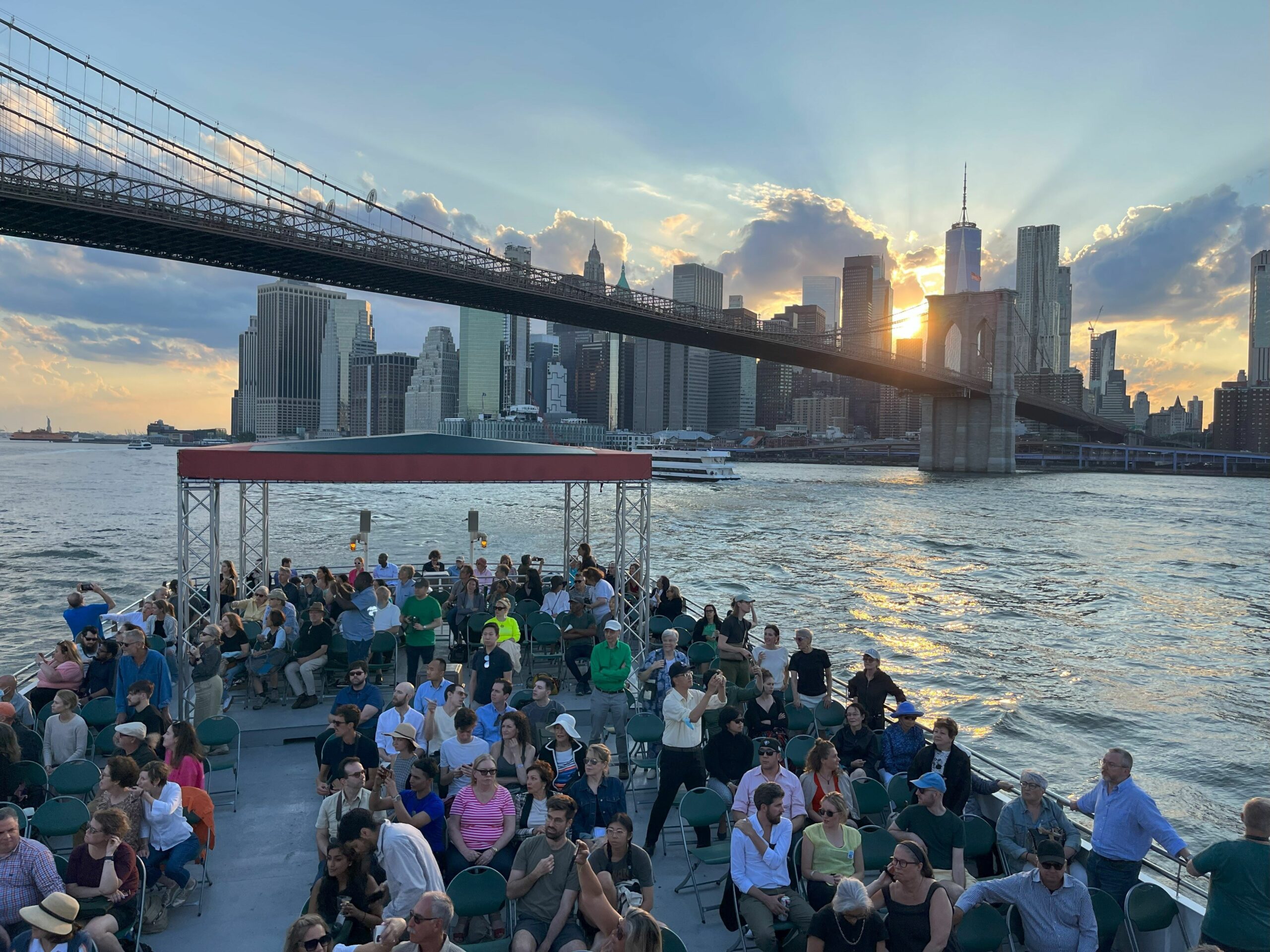 Top deck of a boat in NY harbor, with the Brooklyn Bridge in the background. The sun is about to set over the city skyline.