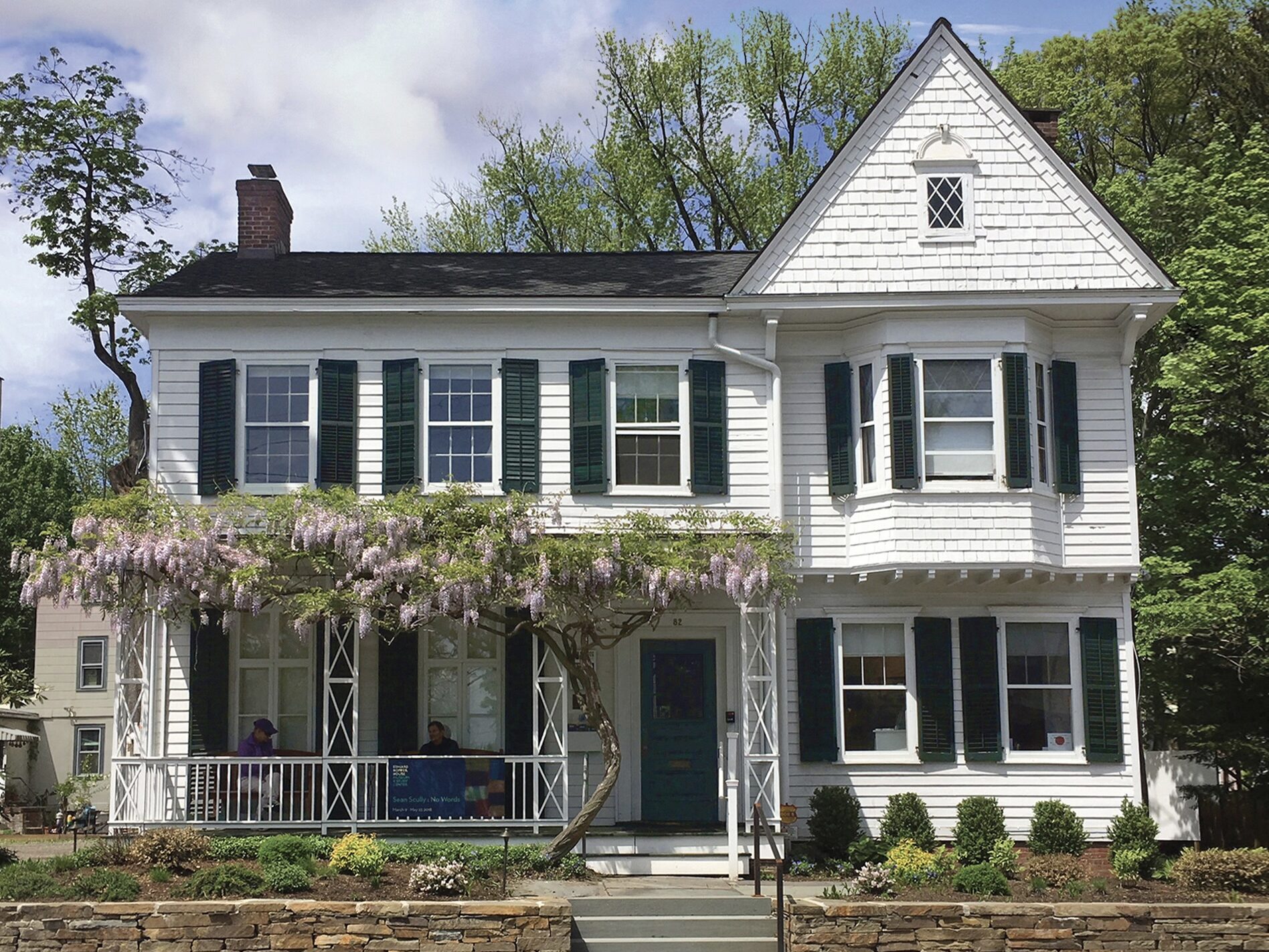 A charming two-story white house with green shutters and a porch . The porch is adorned with lush greenery, and a stone pathway leads to the entrance. Tall trees and bushes surround the house, adding to its quaint and inviting appearance.