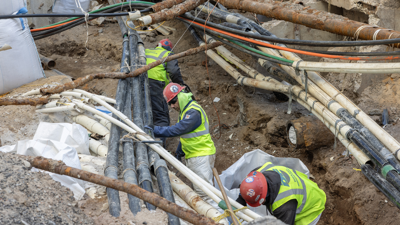 Three men in high-visibility construction vests and hard hats inspect underground water main pipes in a dug out city street.